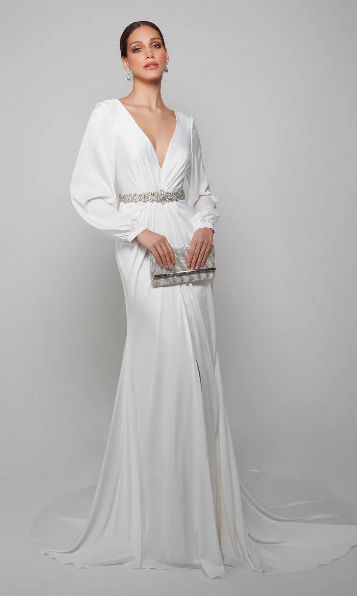 A woman poses against a plain background in the Alyce 7070 Long Formal Dress in Diamond White, featuring a deep V-neck, embellished waistband, long sleeves, front slit, and holding a silver clutch.